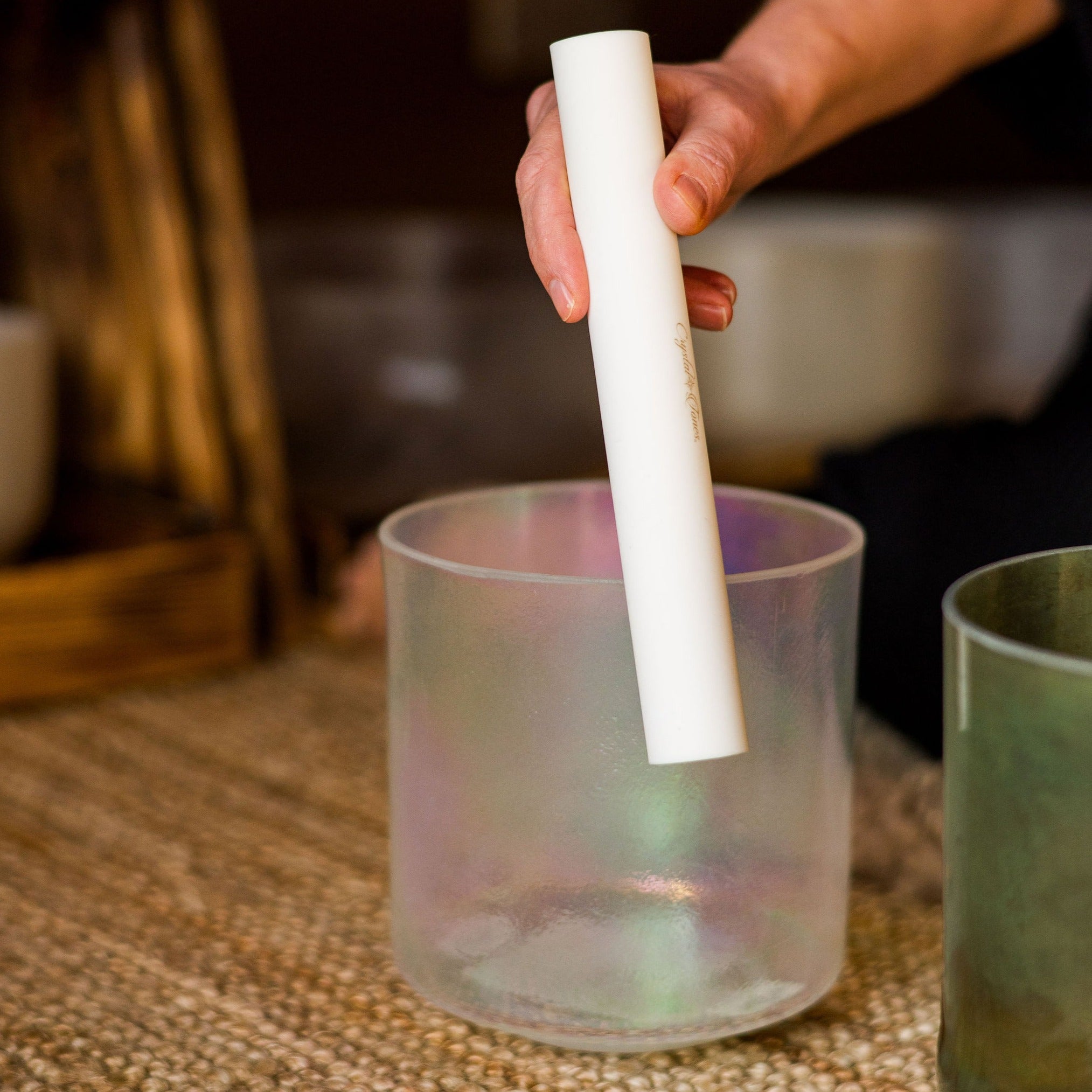 Hand holding a white cylindrical sonic mallet over a crystal sound bowl on a textured surface.