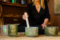 Load image into Gallery viewer, Person using a mallet on a singing bowl in a room with a hessian rug and wooden shelves.
