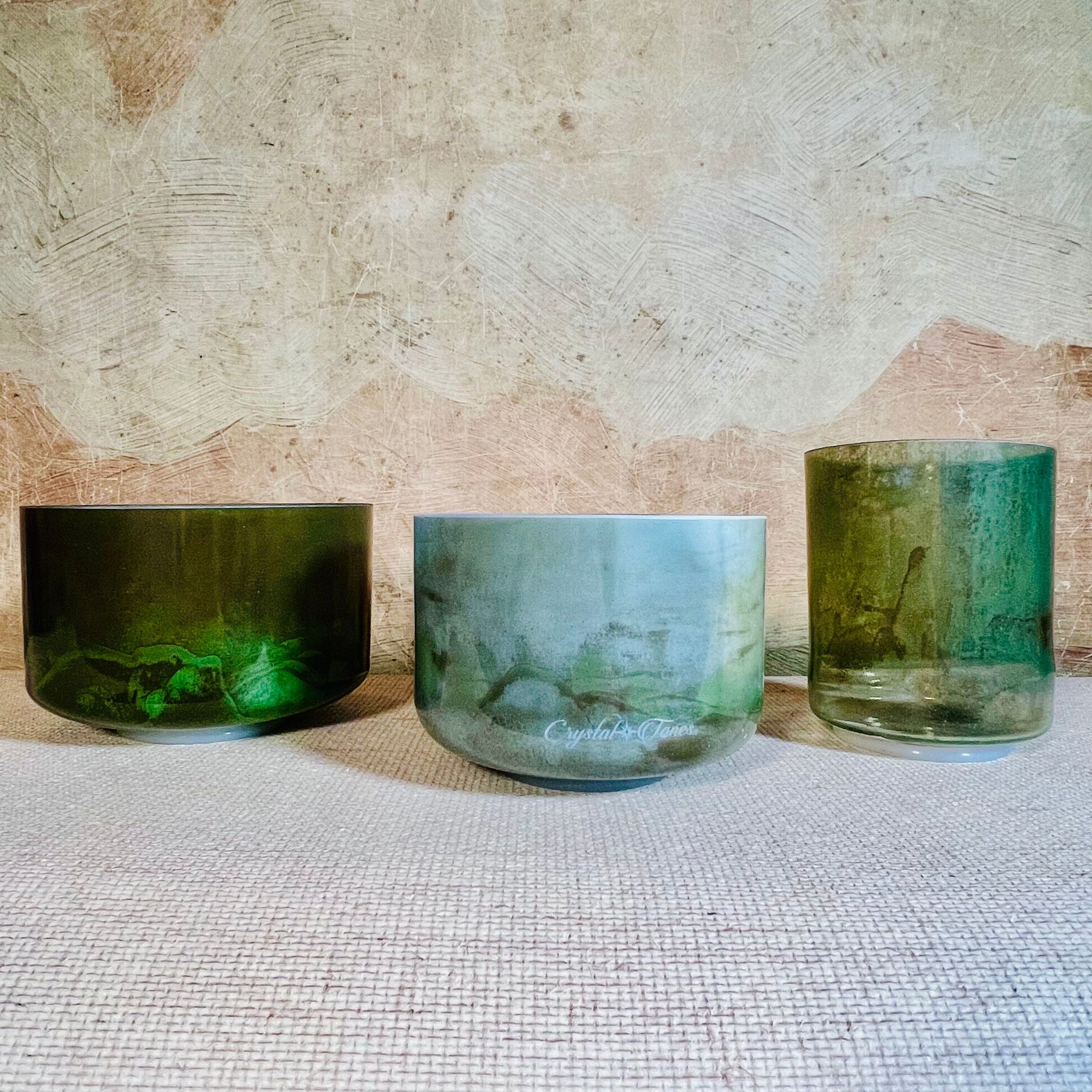 Three green crystal sound bowls of different sizes on a textured surface with a stone wall background.