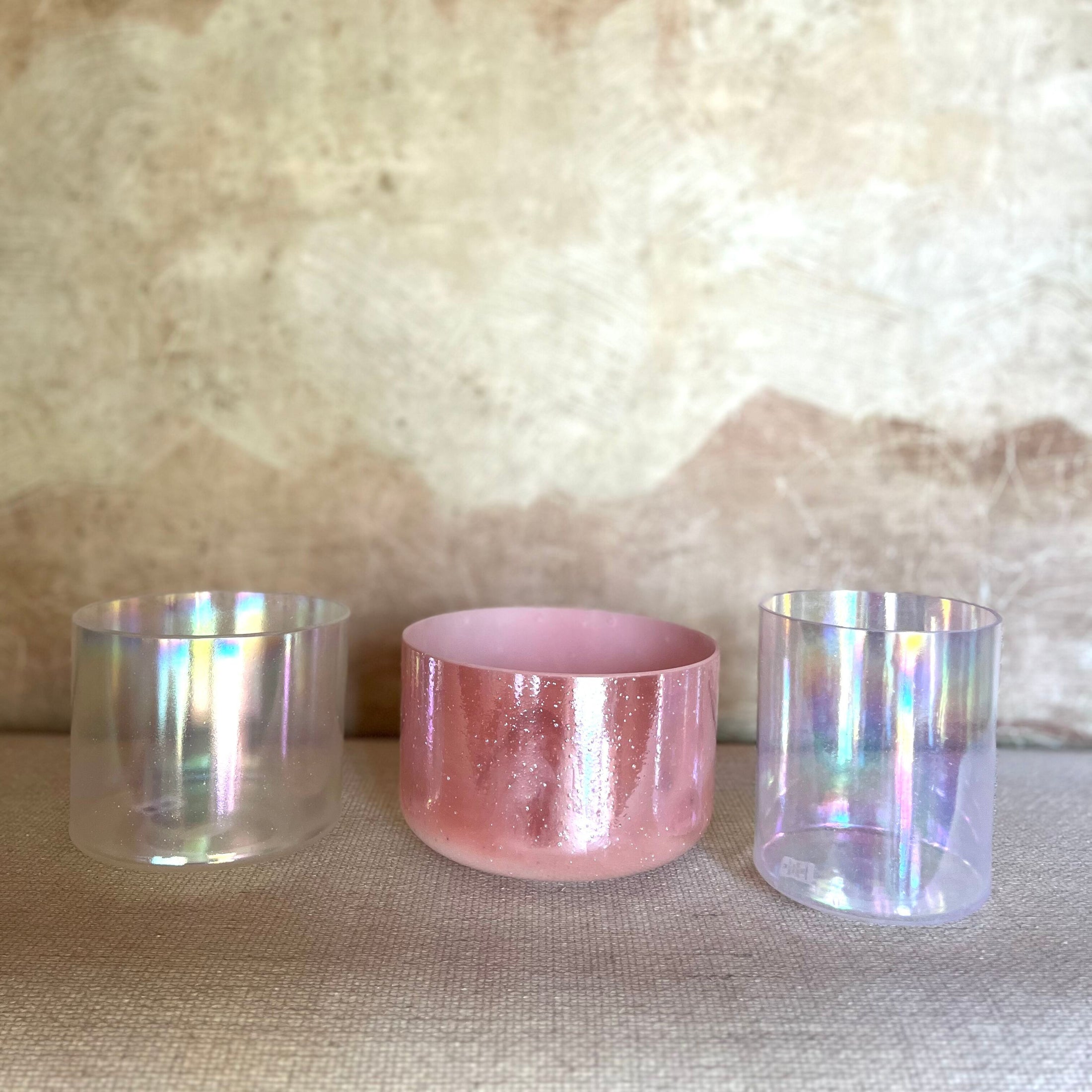 Three crystal sound bowls with iridescent and pink finishes on a textured surface.