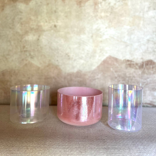 Three crystal sound bowls with iridescent and pink finishes on a textured surface.